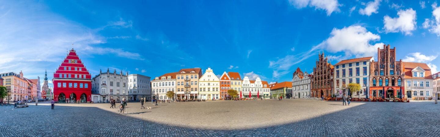 Panoramablick auf einen malerischen europäischen Stadtplatz in Greifswald mit farbenfrohen, historischen Gebäuden in unterschiedlichen Architekturstilen. Links sticht ein leuchtend rotes Gebäude hervor, und Menschen gehen unter einem strahlend blauen Himmel mit vereinzelten Wolken über den Kopfsteinpflasterplatz.