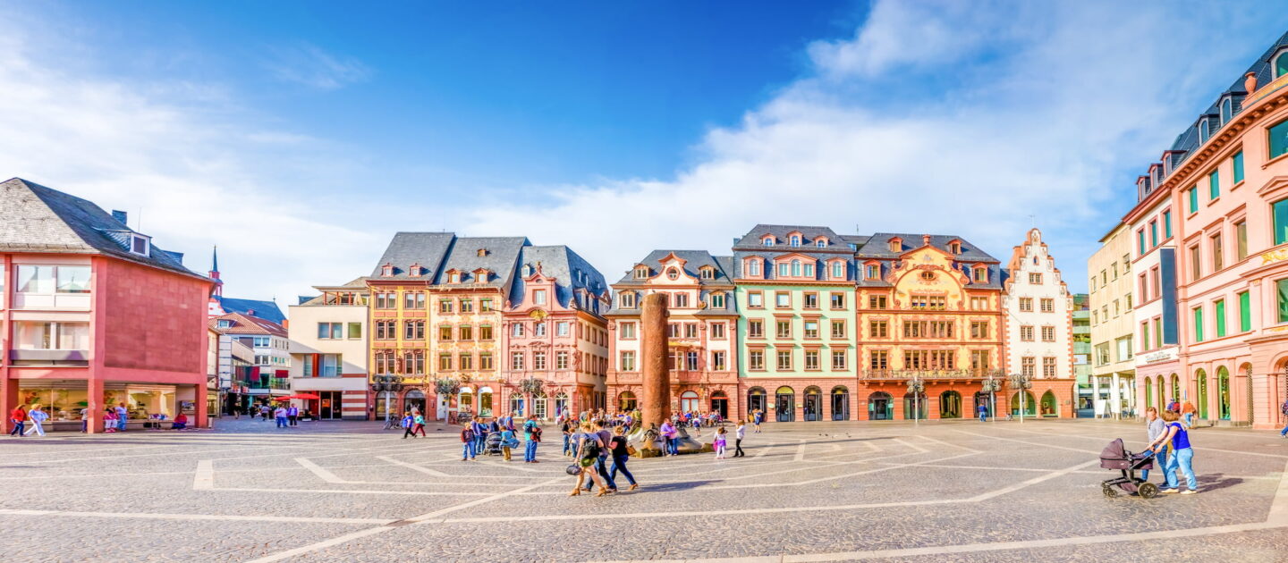 Ein lebhafter Stadtplatz in Mainz zeichnet sich durch farbenfrohe, mehrstöckige Gebäude im europäischen Architekturstil aus. Die Menschen gehen auf dem Kopfsteinpflaster spazieren und unterhalten sich, einige schieben Kinderwagen. Der Himmel ist klar mit ein paar Wolken und die Atmosphäre ist lebhaft und einladend.