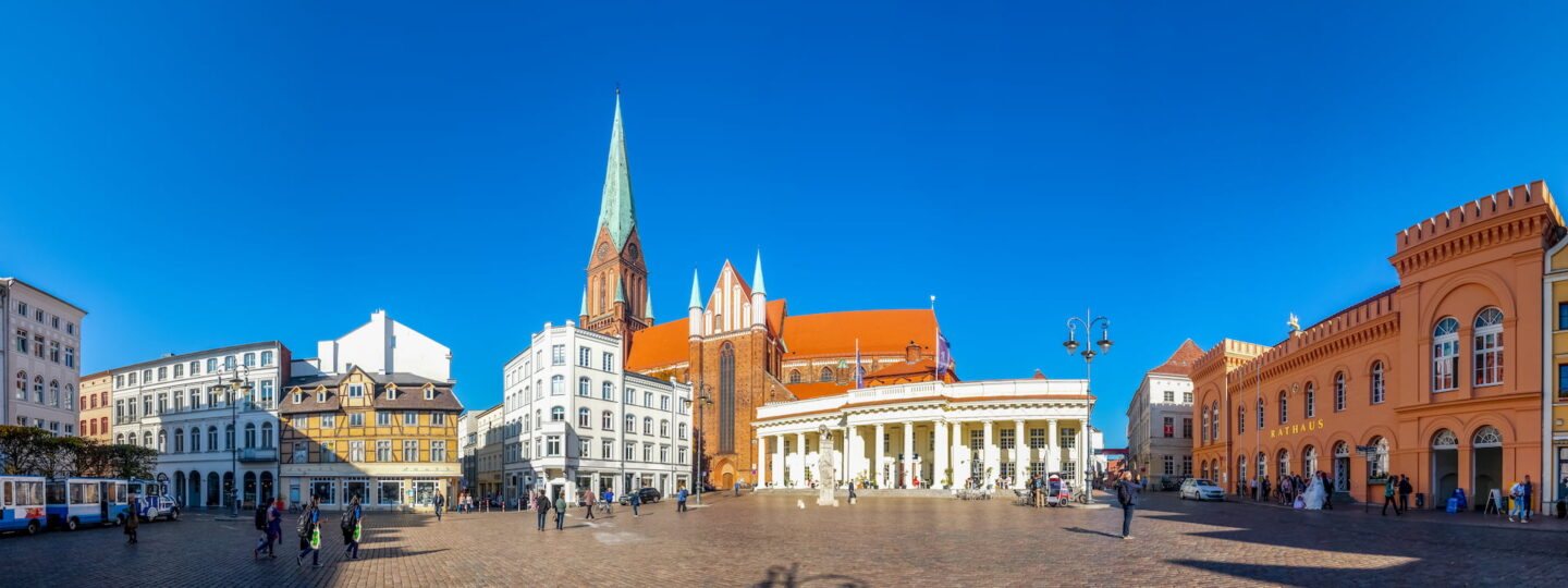 Ein Panoramablick auf den Schweriner Stadtplatz mit historischen Gebäuden unter einem klaren blauen Himmel. Die Szene wird von einer Kirche mit einem hohen, grünen Turm und einer Backsteinfassade dominiert. Um sie herum befinden sich farbenfrohe Gebäude im klassischen europäischen Stil und auf der rechten Seite ein Gebäude aus rotem Backstein.