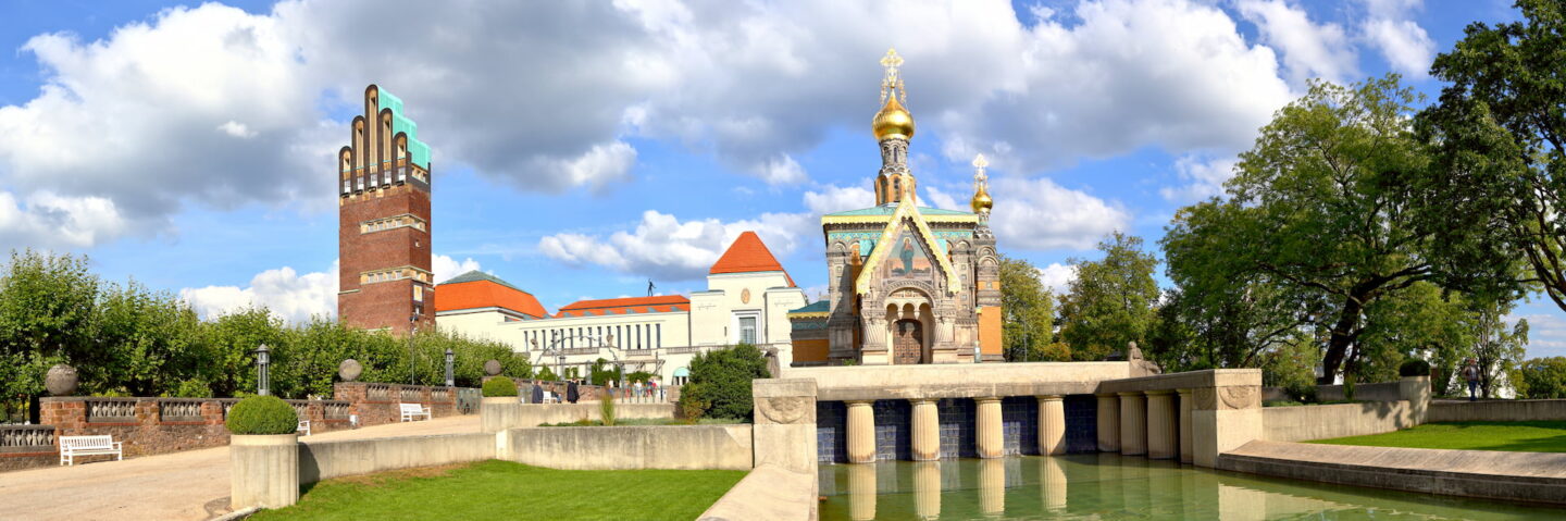 Ein Panoramablick auf die Mathildenhöhe in Darmstadt, Deutschland. Das Bild zeigt die Russische Kapelle mit ihren goldenen Zwiebeltürmen, den markanten Hochzeitsturm mit seinen zahlreichen Bögen, umgebendes Grün und einen klaren blauen Himmel mit vereinzelten Wolken.