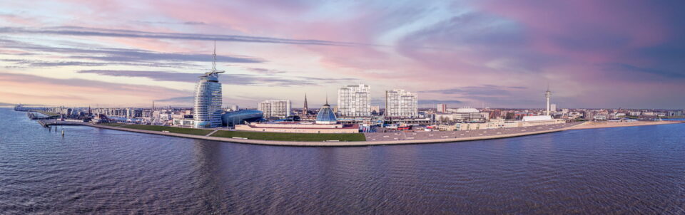 Panoramablick auf die Uferpromenade von Bremerhaven bei Sonnenuntergang. Zu sehen sind mehrere moderne Gebäude entlang der Küste, ein hoher Telekommunikationsturm, eine markante segelförmige Struktur und ein sanfter rosa und violetter Himmel, der sich im ruhigen Wasser spiegelt.