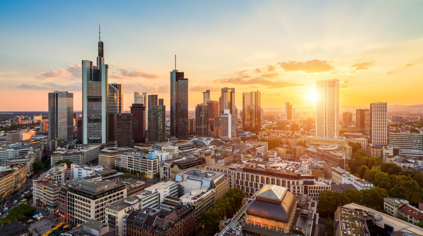 Luftaufnahme der lebendigen Skyline von Frankfurt am Main bei Sonnenuntergang mit modernen Wolkenkratzern und verschiedenen Gebäuden, die in ein warmes goldenes Licht getaucht sind. Die Sonne geht am Horizont unter und wirft ein warmes Licht auf die Stadtlandschaft. Der Himmel ist teilweise bewölkt und weist Orange- und Gelbtöne auf.