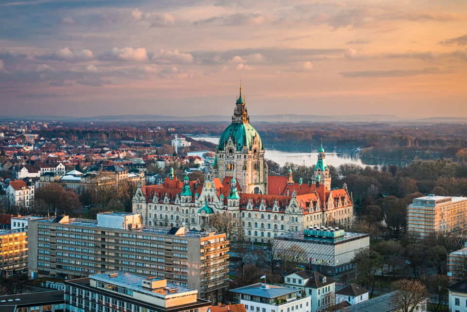 Luftaufnahme des Neuen Rathauses in Hannover bei Sonnenuntergang. Das Gebäude verfügt über eine große Kuppel und Türme, die von Wohn- und Geschäftshäusern umgeben sind. Im Hintergrund sind ein Fluss und eine Mischung aus Bäumen und entfernten Gebäuden unter einem bewölkten Himmel sichtbar.