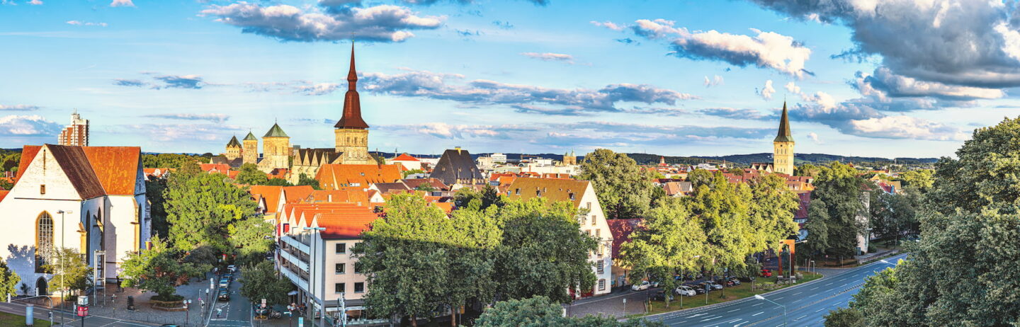 Panoramablick auf die Stadtlandschaft von Osnabrück mit historischen Gebäuden mit roten Ziegeldächern, markanten Kirchtürmen, üppig grünen Bäumen und einer modernen Straße mit Fahrzeugen auf der rechten Seite, alles unter einem blauen Himmel mit vereinzelten Wolken.