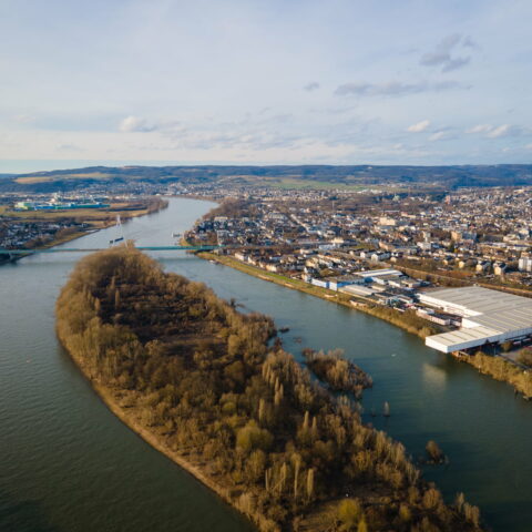 Luftaufnahme eines großen Flusses, der sich durch die dicht besiedelte Stadt Neuwied schlängelt. Im Fluss liegt eine mit Bäumen bewachsene Insel, während Wohnhäuser, Lagerhäuser und Hügel mit vereinzelten Grünflächen das Gebiet unter einem teilweise bewölkten Himmel umgeben.