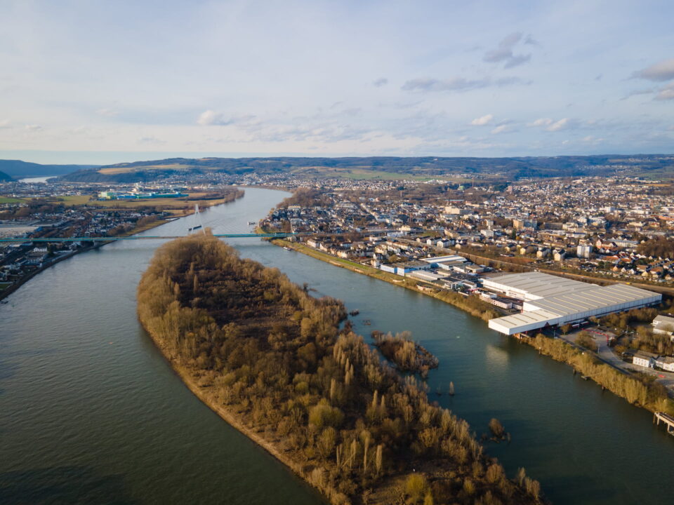 Luftaufnahme eines großen Flusses, der sich durch die dicht besiedelte Stadt Neuwied schlängelt. Im Fluss liegt eine mit Bäumen bewachsene Insel, während Wohnhäuser, Lagerhäuser und Hügel mit vereinzelten Grünflächen das Gebiet unter einem teilweise bewölkten Himmel umgeben.