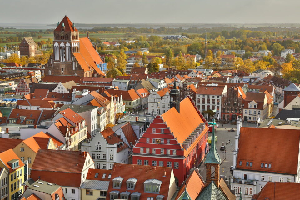 Luftaufnahme der europäischen Stadt Greifswald mit Gebäuden mit roten Dächern und einer markanten Kirche mit hohem Turm. Die Stadt ist von grünen Feldern und Bäumen umgeben und verbindet moderne und historische Architektur. Das Herbstlaub verleiht der Landschaft eine lebendige Farbpalette.