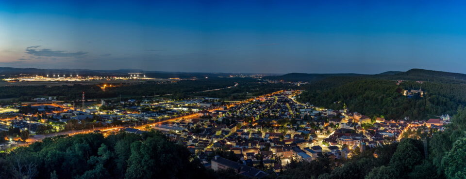 Ein Panoramablick auf Kaiserslautern in der Abenddämmerung mit funkelnden Lichtern von Gebäuden und Straßen. Das Gebiet ist von üppigen, grünen Hügeln umgeben und der Himmel wechselt von Hell- zu Dunkelblau. Die Stadt scheint mit dem fernen Horizont zu verschmelzen.