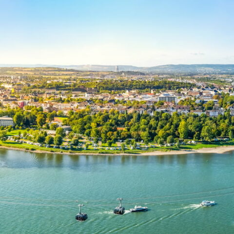 Panoramablick auf eine Stadt entlang einer Flussbiegung mit grünen Landschaften, zahlreichen Gebäuden und mehreren Booten auf dem Wasser. Der Fluss schlängelt sich durch Koblenz, Deutschland, mit üppigem Grün an seinen Ufern und einem hellen, klaren Himmel darüber. Ein perfekter Schnappschuss für jeden Reiseliebhaber.