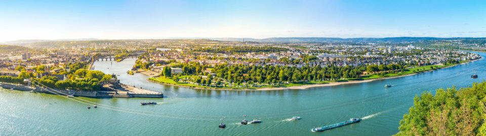 Panoramablick auf eine Stadt entlang einer Flussbiegung mit grünen Landschaften, zahlreichen Gebäuden und mehreren Booten auf dem Wasser. Der Fluss schlängelt sich durch Koblenz, Deutschland, mit üppigem Grün an seinen Ufern und einem hellen, klaren Himmel darüber. Ein perfekter Schnappschuss für jeden Reiseliebhaber.