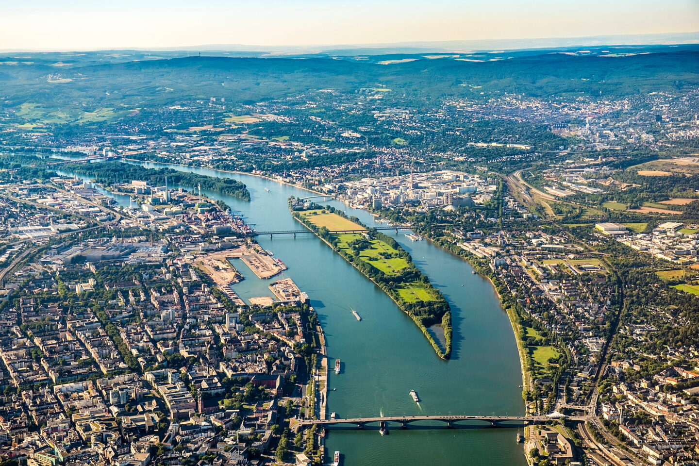 Luftaufnahme von Mainz, einer Stadt, die von einem breiten Fluss durchzogen wird. Mehrere Brücken überspannen den Fluss und verbinden die Stadtgebiete auf beiden Seiten. Gebäude, Straßen und Grünflächen sind sichtbar, in der Ferne sind Hügel und Berge unter einem klaren Himmel zu sehen.