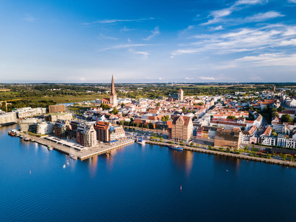 Luftaufnahme der Küstenstadt Rostock mit historischen Gebäuden und Kirchen entlang der Uferpromenade. Das Stadtbild umfasst einen markanten Turm und moderne Gebäude, die von Wasser umgeben sind, vor einer grünen Kulisse und einem teilweise bewölkten blauen Himmel.