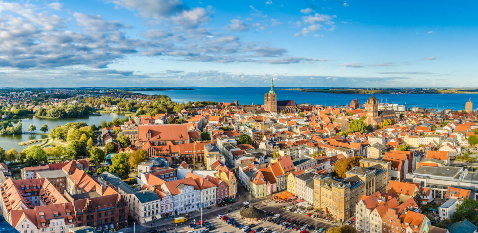 Luftaufnahme der Küstenstadt Stralsund mit ihren rot gedeckten Gebäuden, einer großen Wasserfläche im Hintergrund und einer Mischung aus Grünflächen und Stadtlandschaft unter einem teilweise bewölkten blauen Himmel.