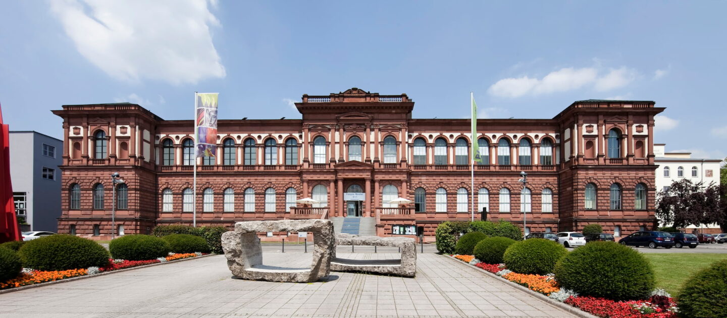 Historisches Museumsgebäude in Kaiserslautern mit einer großen, symmetrischen Fassade mit Bogenfenstern und kunstvollen Details. Im Vordergrund steht eine moderne Skulpturinstallation, umgeben von angelegten Gärten und bunten Blumen. Banner und Flaggen schmücken das Gebäude unter einem teilweise bewölkten Himmel.