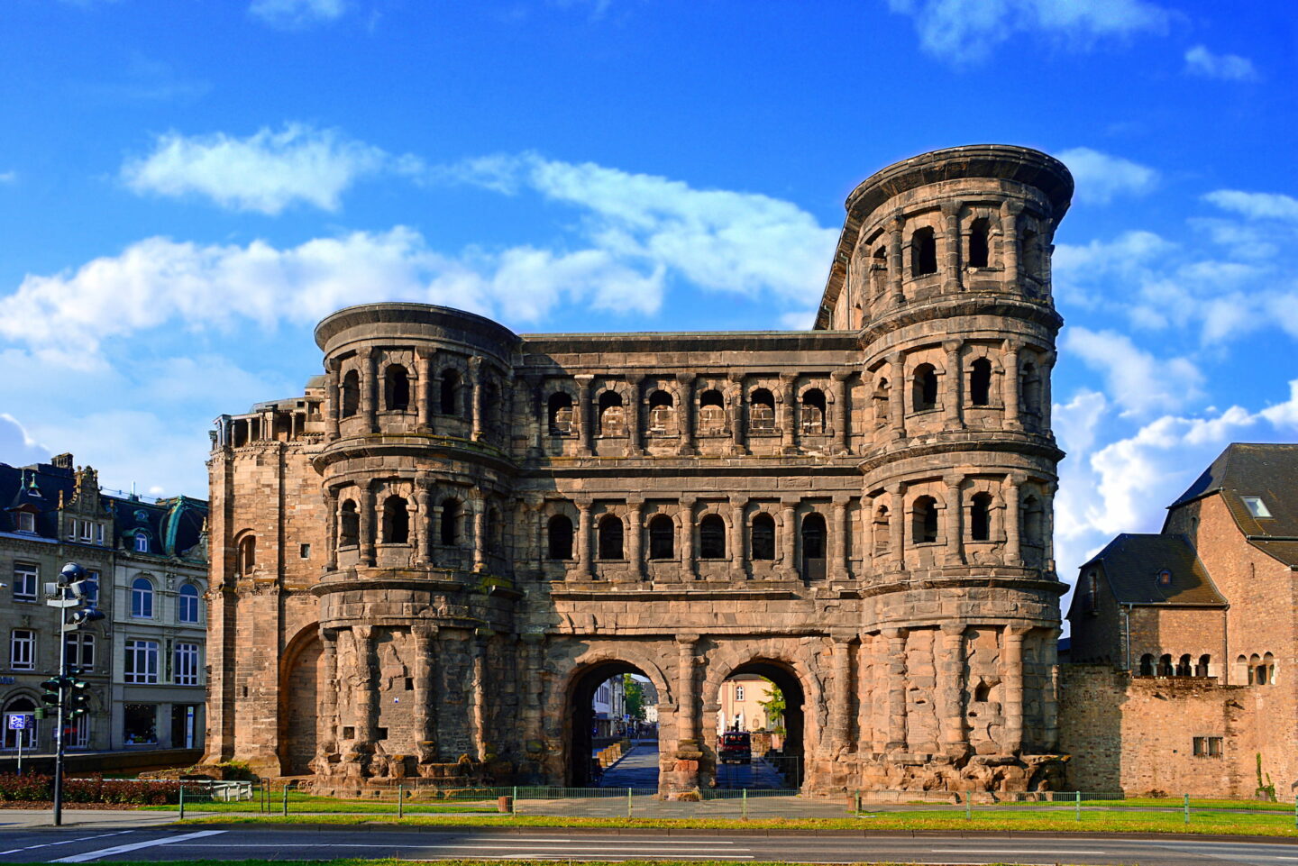 Ein historisches römisches Tor, bekannt als Porta Nigra, ragt hoch vor einem strahlend blauen Himmel mit vereinzelten Wolken in der antiken Stadt Trier in Deutschland empor. Dieses antike Sandsteinbauwerk verfügt über gewölbte Eingänge und mehrere Ebenen, die auf beiden Seiten von runden Türmen flankiert werden. Die umliegenden Gebäude sind teilweise sichtbar.