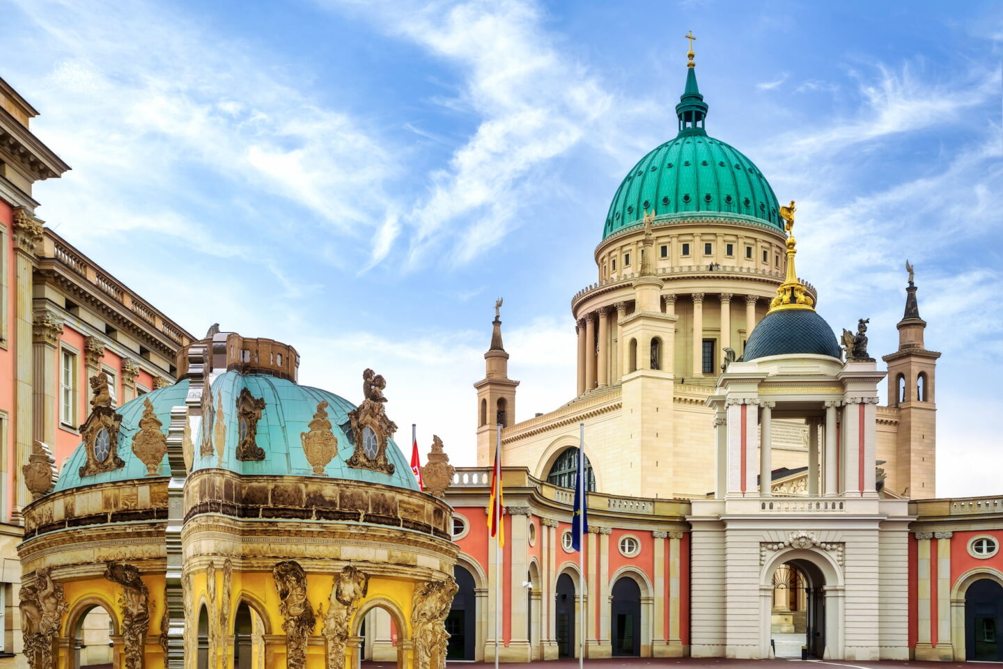 Eine lebendige Architekturszene in Potsdam zeigt ein großes, majestätisches Gebäude mit einer markanten grünen Kuppel, die von einer goldenen Statue gekrönt wird. Flankiert von kleineren Kuppelstrukturen mit aufwendigen Details umfasst die Szene hohe Säulen und farbenfrohe Fassaden unter einem klaren blauen Himmel.