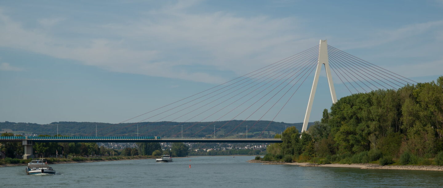 Ein breiter Fluss mit mehreren Booten wird von einer Schrägseilbrücke überspannt. Die Neuwied-Brücke verfügt über einen einzelnen, hohen Turm, von dem aus zahlreiche Kabel diagonal verlaufen, um das Brückendeck zu stützen. Die Flussufer sind von dichtem Grün gesäumt und in der Ferne sind Hügel unter einem teilweise bewölkten Himmel sichtbar.