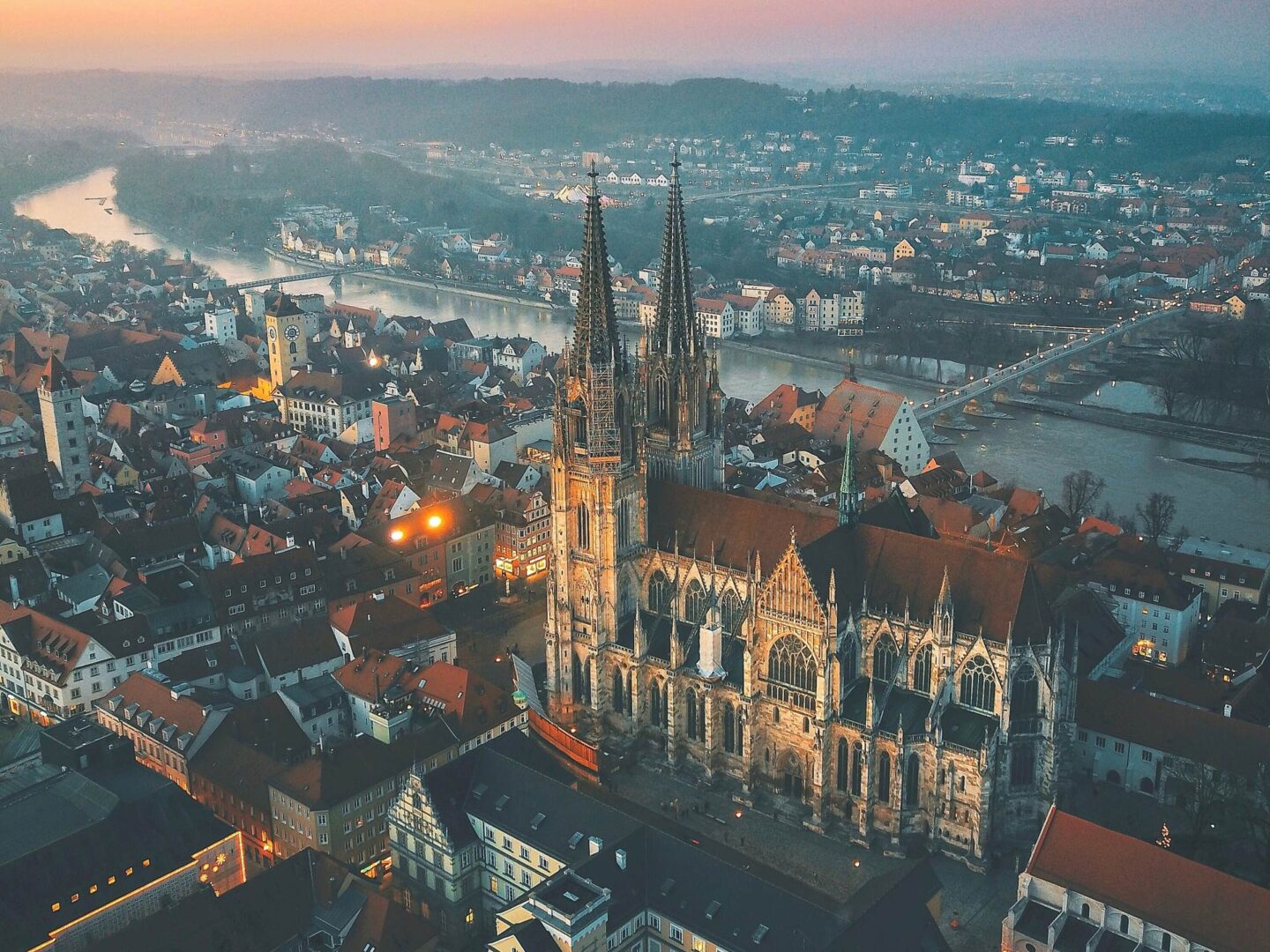 Luftaufnahme der historischen europäischen Stadt Regensburg bei Sonnenuntergang mit einer markanten gotischen Kathedrale mit zwei hohen Türmen. Die Stadt liegt an einem gewundenen Fluss und hat zahlreiche Gebäude und enge Straßen, alles umgeben von malerischen Hügeln und einem sanften Abendhimmel – ein ideales Reiseziel in Deutschland.