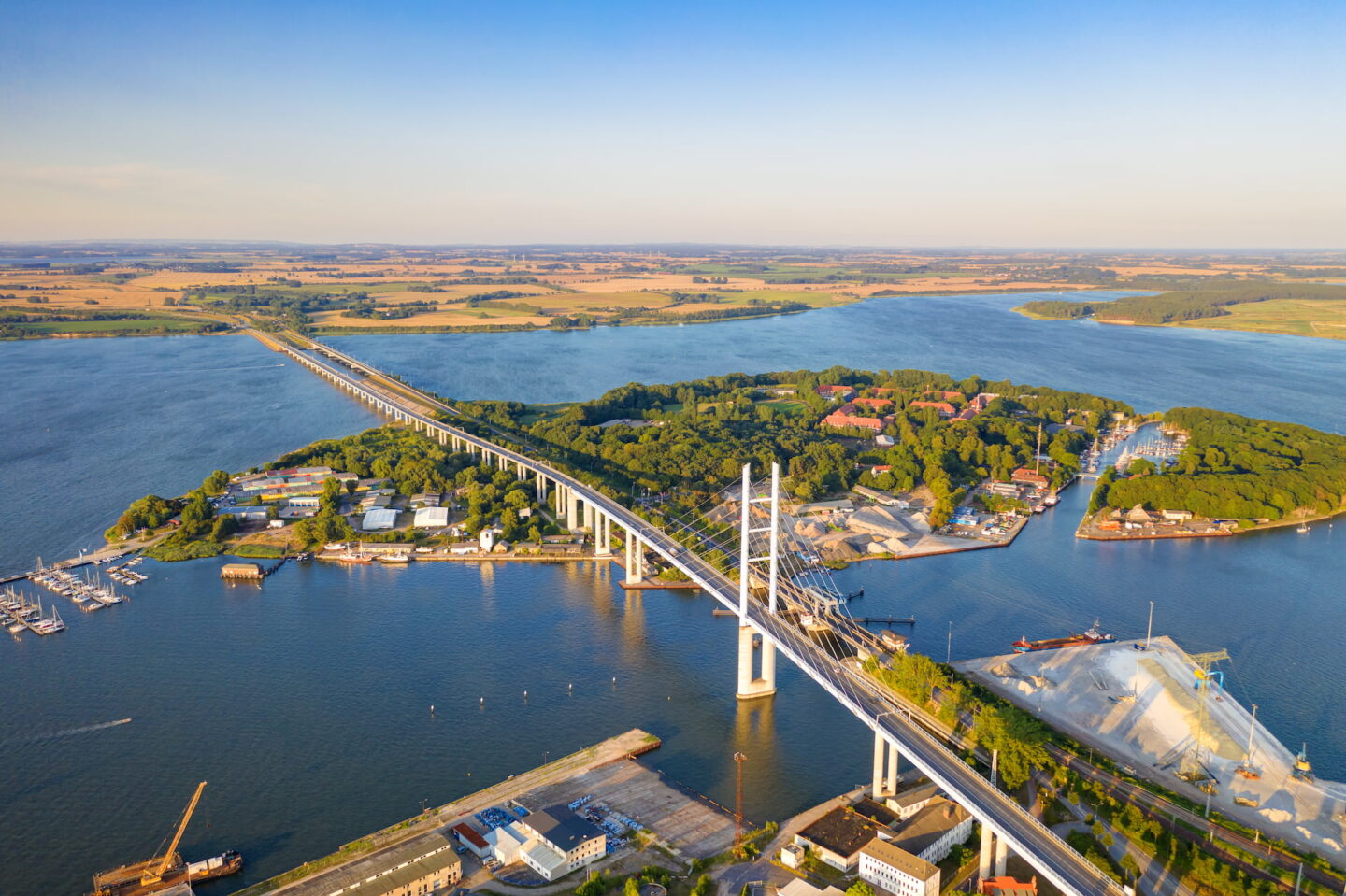 Luftaufnahme einer langen Brücke, die zwei Landmassen über ein Gewässer, möglicherweise die Ostsee, verbindet. In der Umgebung sind üppige grüne Landschaften, Industriegebäude und Boote zu sehen, die am Ufer in der Nähe der historischen Stadt Stralsund angedockt sind. Der Himmel ist klar und warmes Sonnenlicht erhellt die Szene.