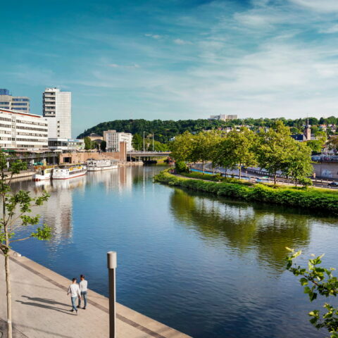 Eine malerische Stadtlandschaft am Flussufer in Saarbrücken mit modernen Gebäuden, einem Spazierweg und Bäumen am Wasser. Zwei Menschen gehen den Weg entlang, während ein Boot am Flussufer angedockt ist. Der Himmel ist klar mit ein paar Wolken und im Hintergrund ist üppiges Grün zu sehen.