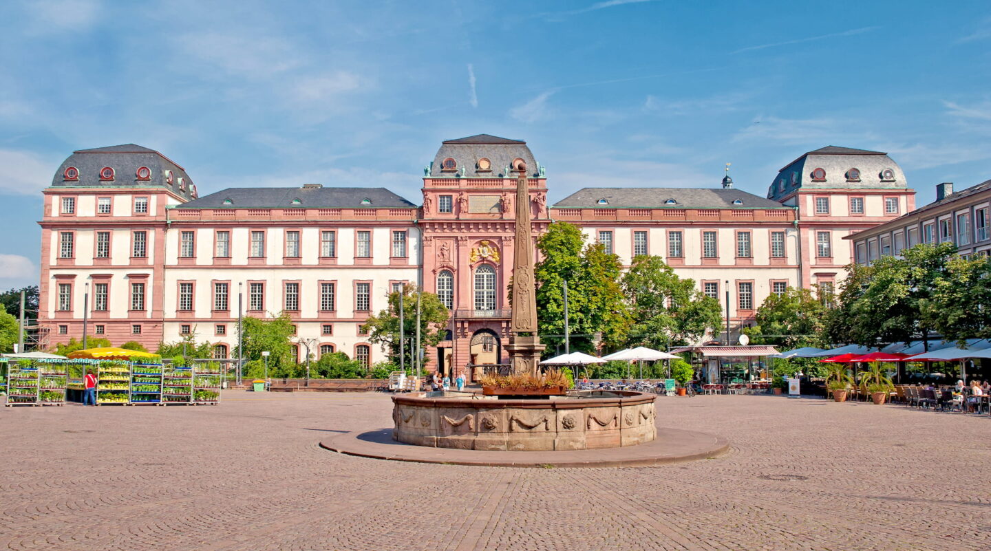 Ein großes, reich verziertes historisches Gebäude mit einer rosa-weißen Fassade, die an den architektonischen Charme von Darmstadt erinnert, weist im Hintergrund einen zentralen Turm auf. Im Vordergrund ziert ein runder Steinbrunnen den geräumigen, gepflasterten Platz. Rechts tragen Straßencafés mit Sonnenschirmen und Tischen zur lebhaften Szene bei.