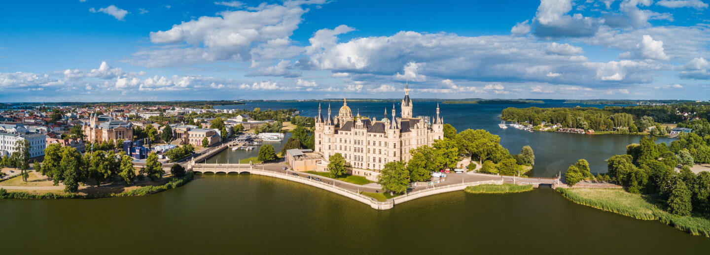 Luftaufnahme des Schweriner Schlosses, das majestätisch auf einer Insel im Schweriner See liegt. Umgeben von üppigem Grün und Wasser besticht das historische Gebäude durch kunstvolle Architektur mit Türmen. Im Hintergrund sind die Stadt Schwerin und weitere Gewässer unter einem teilweise bewölkten Himmel zu sehen.