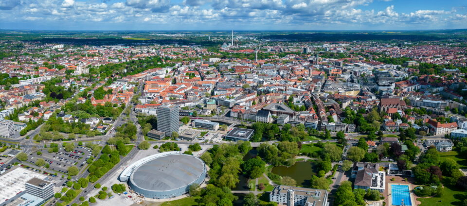 Luftaufnahme von Braunschweig mit einer Mischung aus modernen und historischen Gebäuden, umgeben von grünen Parks und Bäumen. Im Vordergrund ist ein großes kreisförmiges Gebäude zu sehen, in der Nähe befindet sich ein kleineres Gewässer. Die Stadt erstreckt sich unter einem teilweise bewölkten Himmel bis zum Horizont.