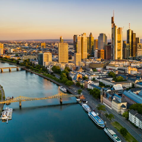 Luftaufnahme von Frankfurt bei Sonnenuntergang. Die Skyline wird von modernen Wolkenkratzern dominiert, der Main fließt anmutig durch die Stadt. Eine malerische Brücke überquert den Fluss und Grünflächen umgeben die Stadtlandschaft. Die Szene ist in warmes, goldenes Licht getaucht.