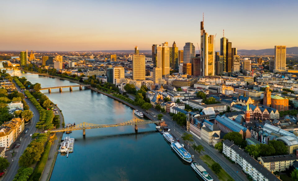 Luftaufnahme von Frankfurt bei Sonnenuntergang. Die Skyline wird von modernen Wolkenkratzern dominiert, der Main fließt anmutig durch die Stadt. Eine malerische Brücke überquert den Fluss und Grünflächen umgeben die Stadtlandschaft. Die Szene ist in warmes, goldenes Licht getaucht.