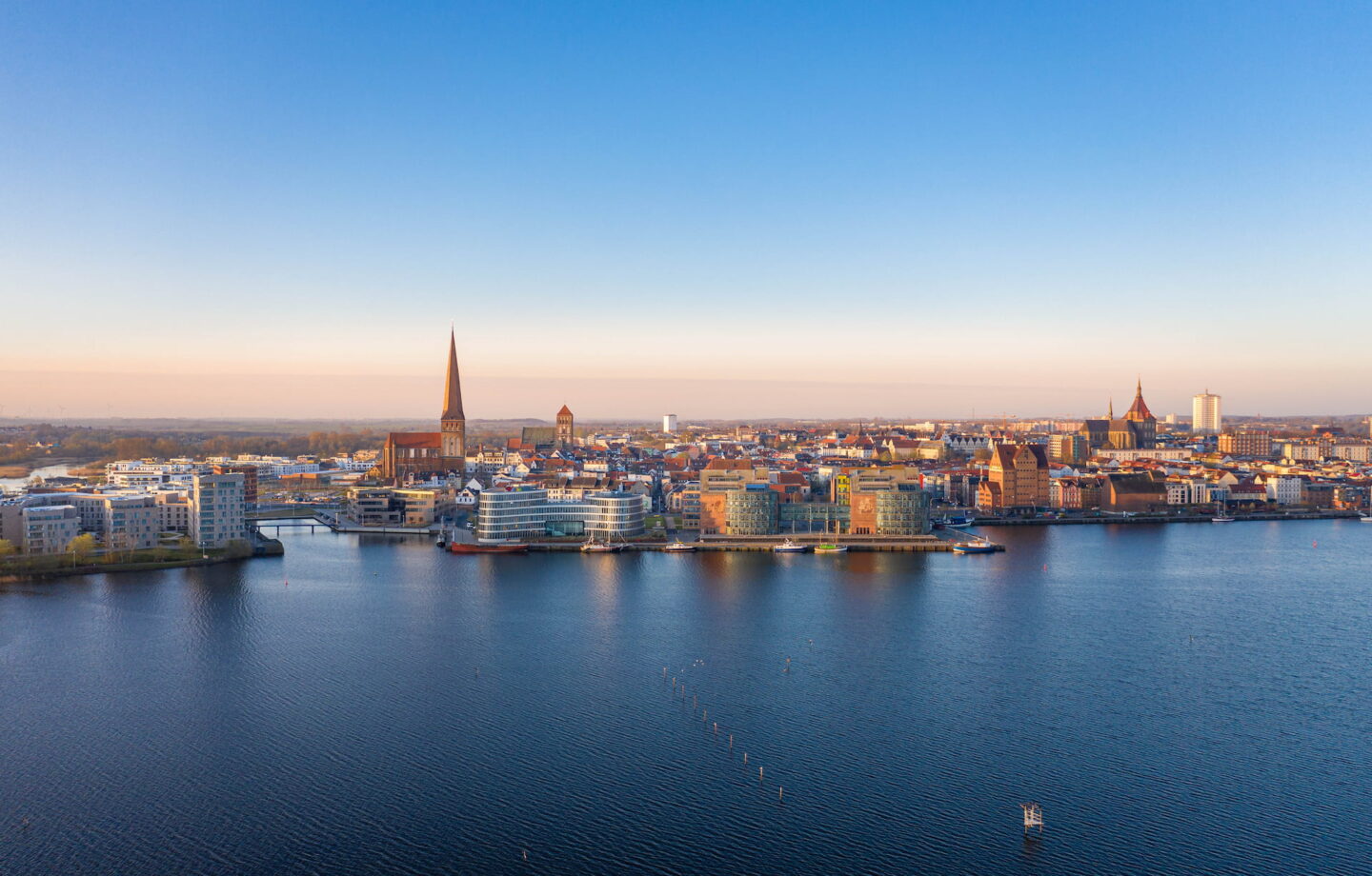 Ein Panoramablick auf Rostock im Morgen- oder Abendgrauen mit einem ruhigen blauen Gewässer im Vordergrund. Die Skyline der Stadt umfasst moderne Gebäude, historische Bauten und mehrere spitze Kirchtürme unter einem klaren, von Blau nach Orange verlaufenden Himmel.