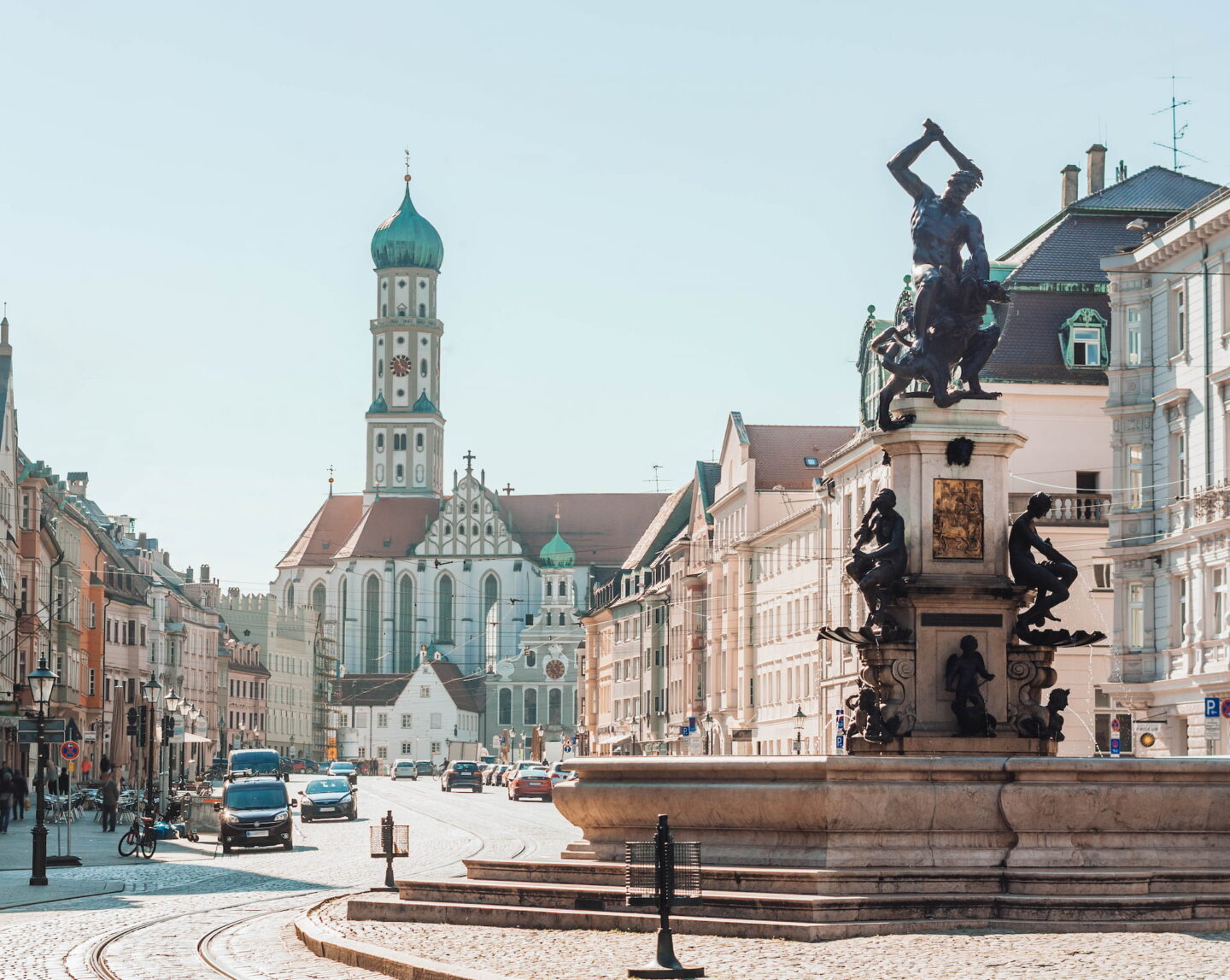 Eine malerische europäische Stadtstraße in Augsburg mit einer historischen Kirche mit einer grünen Kuppel im Hintergrund. Im Vordergrund ist ein detailreiches Statuendenkmal zu sehen, das von Gebäuden umgeben ist. Auf der Straße sind ein paar Autos und Menschen, die einen sonnigen Tag genießen.