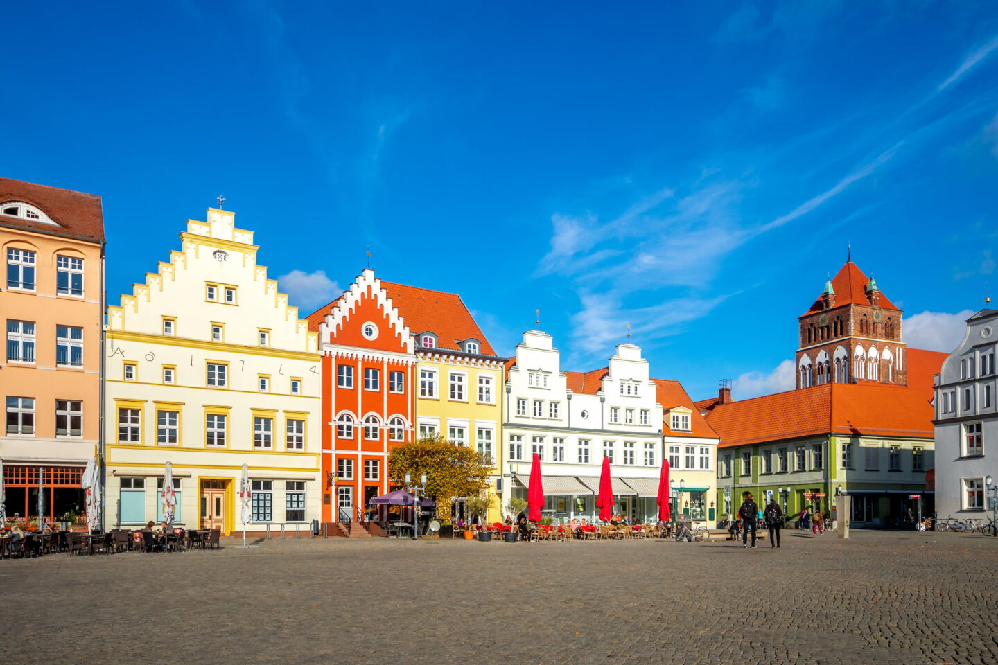 Ein malerischer Stadtplatz in Greifswald mit farbenfrohen, historischen Gebäuden mit roten, gelben, weißen und grünen Fassaden. Der Platz ist gesäumt von Straßencafés und ein paar Passanten. Der Himmel ist klar und blau.