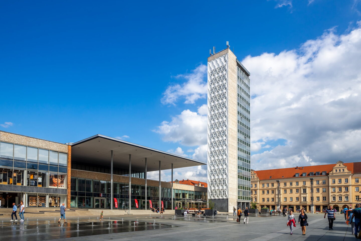 Auf einem großen Platz in Neubrandenburg steht ein modernes Hochhaus mit weißer, kunstvoller Fassade und Glasfenstern. Daneben steht ein Flachbau mit großem Überhang und Glasfront. An diesem sonnigen Tag mit vereinzelten Wolken laufen nur wenige Menschen auf dem Platz herum.