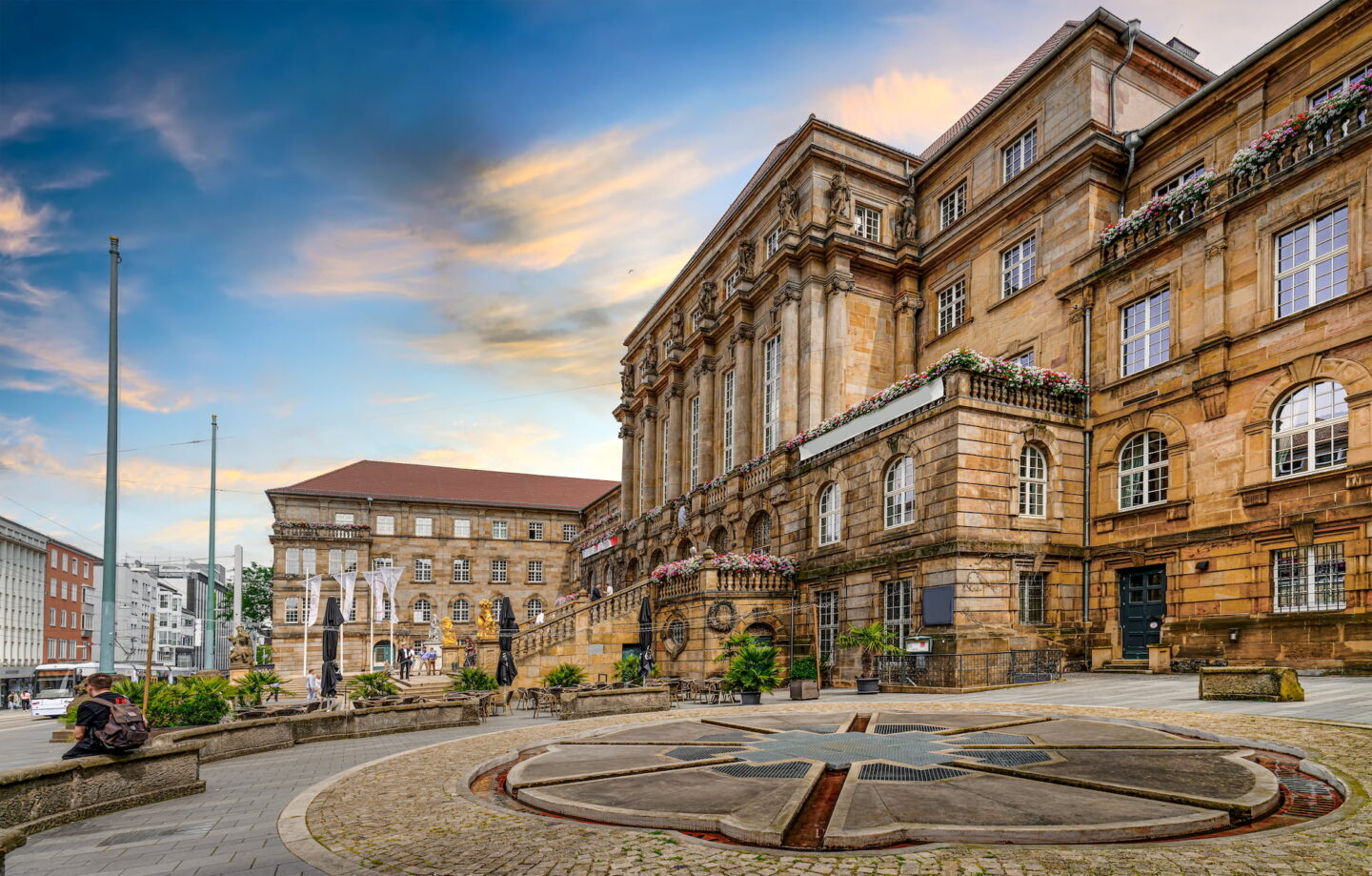 Ein historisches, reich verziertes Gebäude in Kassel ist mit großartigen architektonischen Details, großen Säulen und Blumendekorationen unter einem teilweise bewölkten Himmel bei Sonnenuntergang zu sehen. Im Vordergrund ist ein kreisförmiger Steinplatz mit sternförmigem Design zu sehen, umgeben von städtischen Gebäuden.