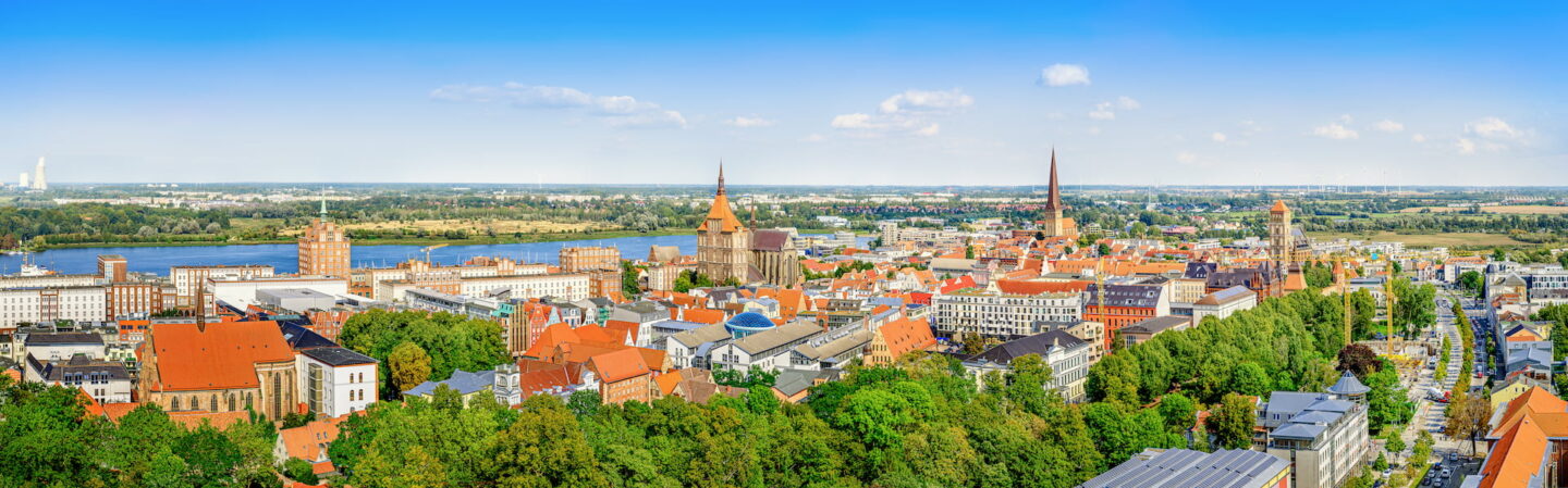 Panoramablick auf Rostock mit seinen rot gedeckten Gebäuden, mehreren hohen Türmen und einem breiten Fluss, der daran entlang fließt. Die Szene umfasst historische Architektur, üppige grüne Bäume und eine weitläufige Landschaft, die sich unter einem klaren blauen Himmel in die Ferne erstreckt.