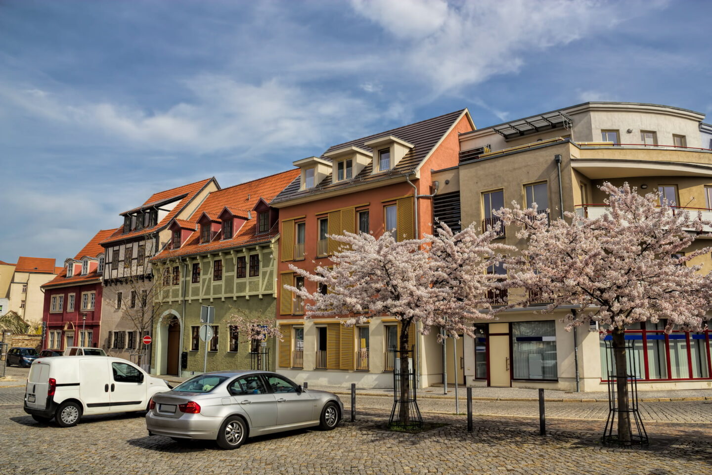 Eine charmante Straße in Aschersleben ist gesäumt von farbenfrohen Gebäuden unter einem teilweise bewölkten Himmel. Im Vordergrund sind drei blühende Kirschbäume und mehrere geparkte Autos zu sehen, darunter ein weißer Lieferwagen und eine silberne Limousine. Die Umgebung hat eine malerische Atmosphäre.