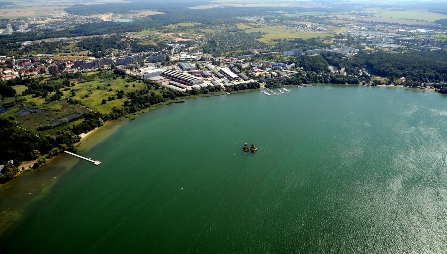 Luftaufnahme des großen Neubrandenburger Sees mit dicht bewaldetem Ufer. In der Mitte des Sees befindet sich eine kleine Insel, von der linken Seite ragt ein Steg ins Wasser. In Ufernähe und im Hintergrund sind zahlreiche Gebäude und ein Wohngebiet zu erkennen.