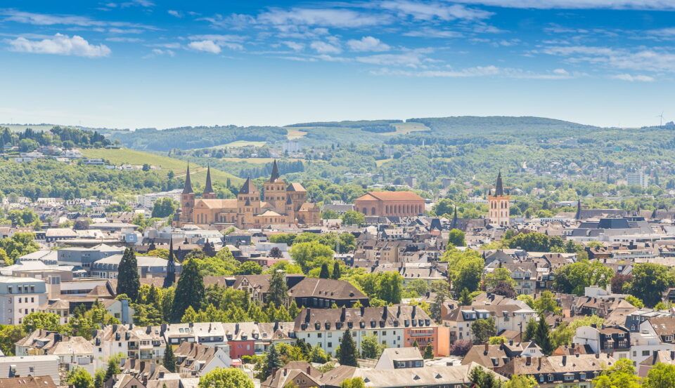 Luftaufnahme von Trier mit einer Mischung aus historischen und modernen Gebäuden, darunter mehrere markante Kirchtürme. Die Landschaft zeichnet sich durch üppiges Grün und sanfte Hügel im Hintergrund unter einem strahlend blauen Himmel mit vereinzelten Wolken aus.