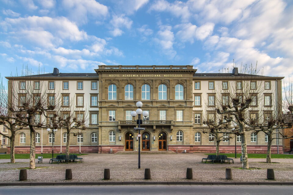 Vorderansicht eines großen, historischen Gebäudes mit drei Stockwerken und einer Steinfassade mit der Aufschrift „Muzeum Akademia Lubowiczska w Rogowicach“. Der Hof vor dem Gebäude in Gießen besteht aus kahlen Bäumen, Bänken und sorgfältig angeordneten Laternenpfählen.