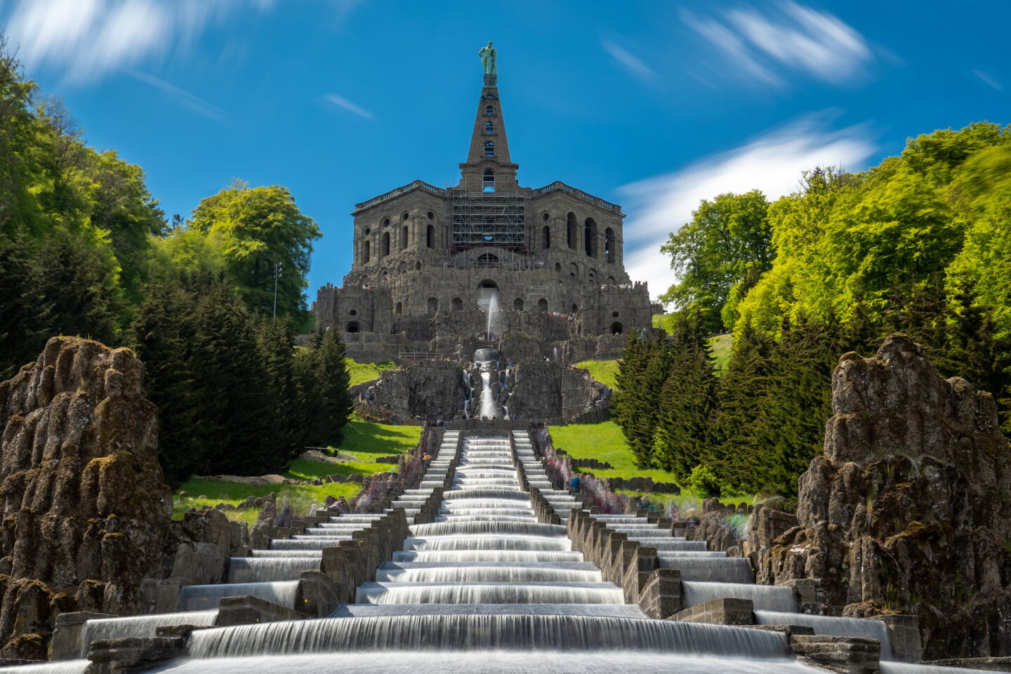 Ein Bild des Herkules-Denkmals in Kassel, Deutschland, mit kaskadierenden Wasserfällen im Vordergrund. Das Denkmal verfügt über eine massive, gestufte Treppe, die zu einem Bauwerk führt, auf dem eine Herkules-Statue steht, umgeben von üppigem Grün und unter Kassels strahlend blauem Himmel.