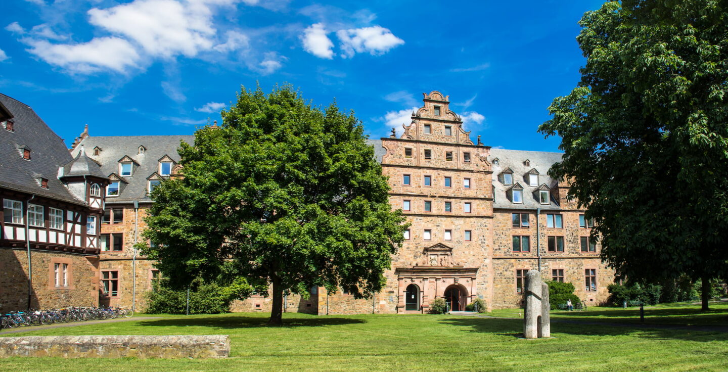 Ein großes, historisches Gebäude in Gießen mit einer detailreichen Fassade ist von üppigem Grün und Bäumen unter einem strahlend blauen Himmel mit vereinzelten weißen Wolken umgeben. Das Gebäude verfügt über zahlreiche Fenster und Dachgauben und im Vordergrund steht eine Steinskulptur.