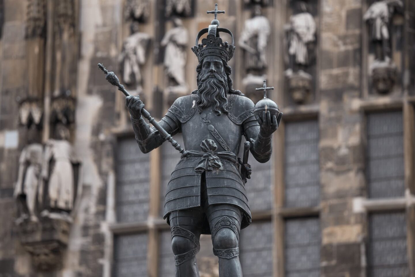 Eine Statue eines mittelalterlichen Königs in voller Rüstung mit Krone und Umhang steht stolz in der historischen Stadt Aachen. In seiner rechten Hand hält er ein Zepter und in seiner linken einen Reichsapfel. Im Hintergrund ist alte Steinarchitektur mit Fensterscheiben und geschnitzten Figuren zu sehen.