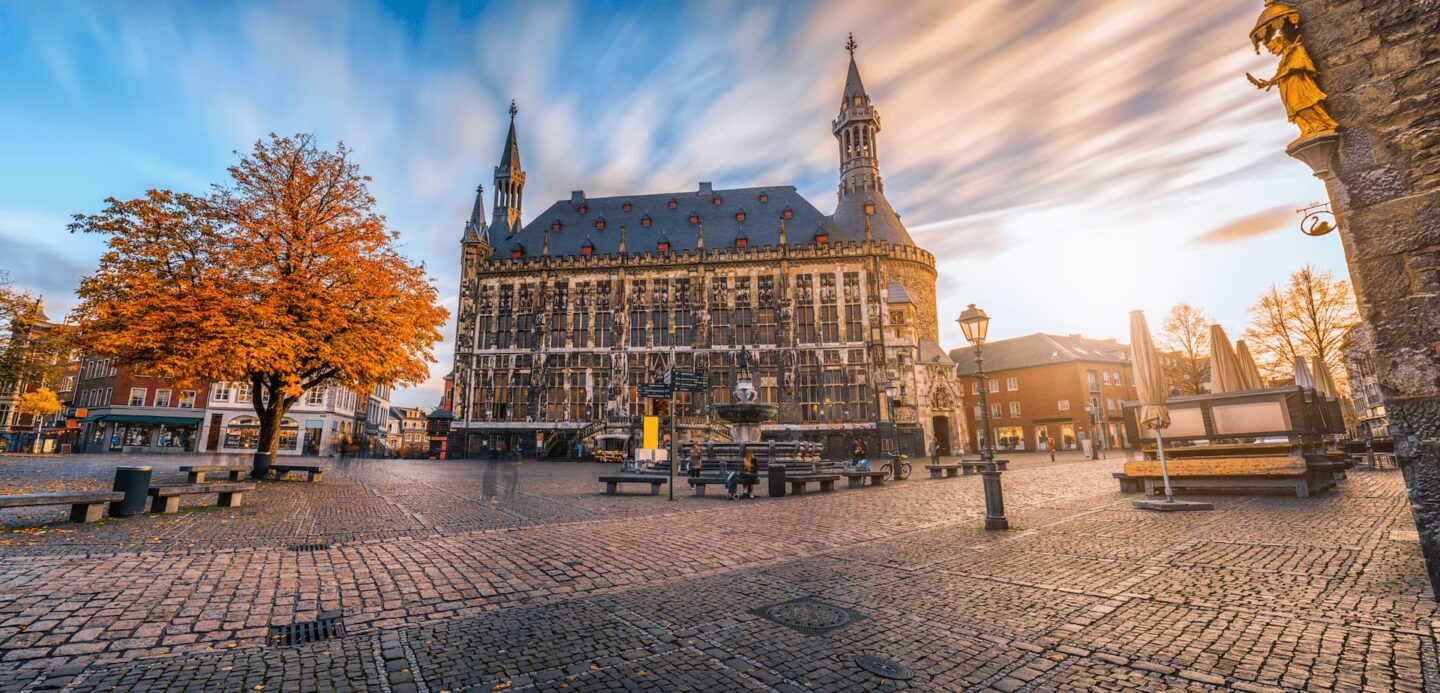 Ein malerischer Stadtplatz in Aachen mit einem historischen Gebäude im gotischen Stil mit kunstvoller Architektur und Türmen, vor einem leuchtenden, wolkigen Himmel bei Sonnenuntergang. Der Platz hat Kopfsteinpflaster, einen einsamen Herbstbaum, Bänke und eine Statue auf der rechten Seite, was ihn perfekt für den Tourismus in Deutschland macht.