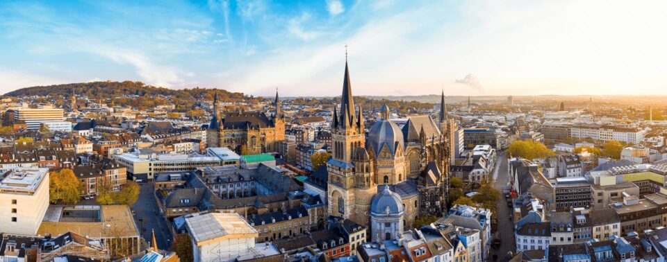 Ein Panoramablick auf die historische europäische Stadt Aachen zeigt einen markanten Dom mit mehreren Türmen und Kuppeln. Das Stadtbild umfasst eine Mischung aus alten und modernen Gebäuden, mit einer Kulisse aus fernen Bergen unter einem klaren, blauen Himmel.