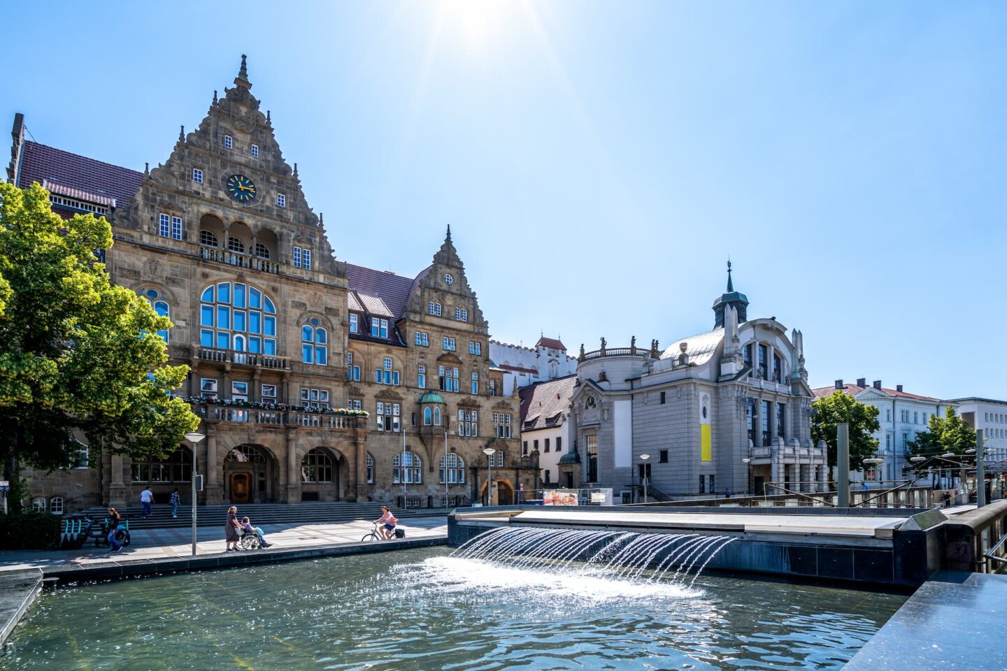 Ein historischer Stadtplatz in Bielefeld mit einem Brunnen im Vordergrund, flankiert von einem prachtvollen, kunstvollen Gebäude mit großen Bogenfenstern und einer Uhr auf der einen Seite und einem kleineren, eleganten Gebäude auf der anderen Seite. In der Nähe sitzen und gehen Menschen unter klarem blauen Himmel spazieren.