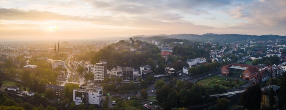 Ein Panoramablick auf die Stadtlandschaft Bielefelds bei Sonnenuntergang umfasst eine Mischung aus modernen und historischen Gebäuden, üppigen grünen Bäumen und gewundenen Straßen. Der Himmel ist mit dem sanften, goldenen Licht der untergehenden Sonne erfüllt und schafft eine heitere und malerische Atmosphäre.