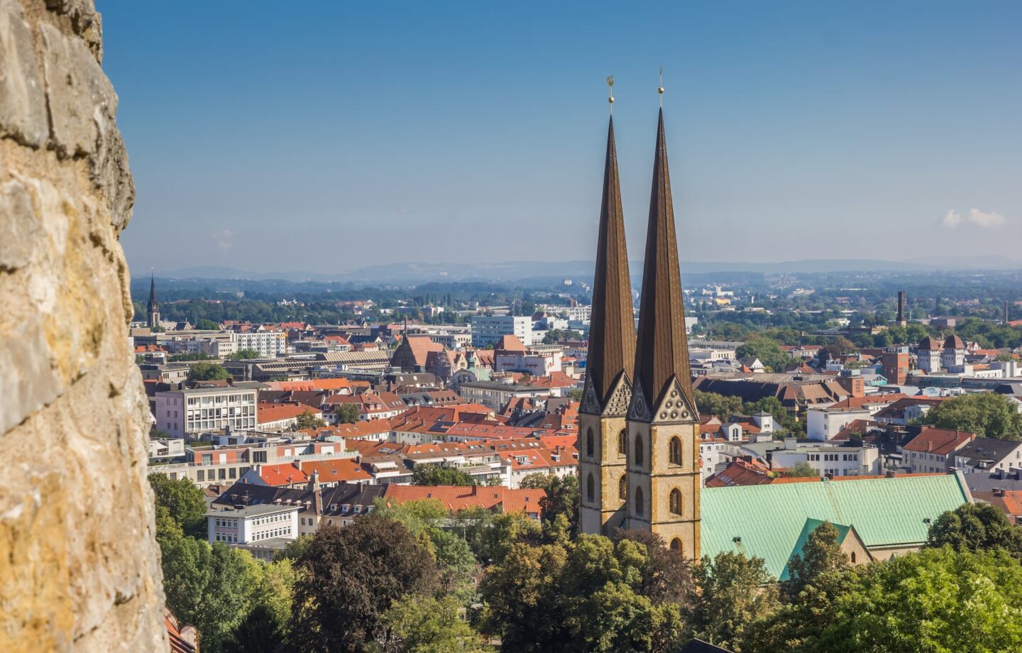 Ein Panoramablick auf die Stadtlandschaft Bielefelds zeigt im Vordergrund zwei hohe, spitze Kirchtürme mit braunen Dächern. Die Umgebung ist geprägt von einer Mischung aus historischen und modernen Gebäuden, Häusern mit roten Dächern und üppigem Grün unter einem strahlend blauen Himmel.