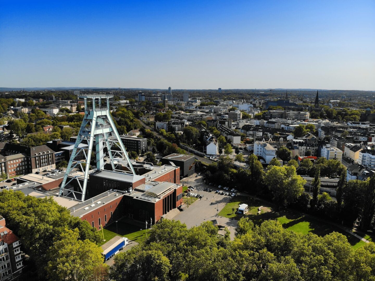 Luftaufnahme von Bochum mit einem markanten Bergbauturm, der von verschiedenen Gebäuden umgeben ist. Die Umgebung ist üppig mit grünen Bäumen und offenen Flächen, und der Horizont zeigt eine ausgedehnte Skyline unter einem klaren blauen Himmel.