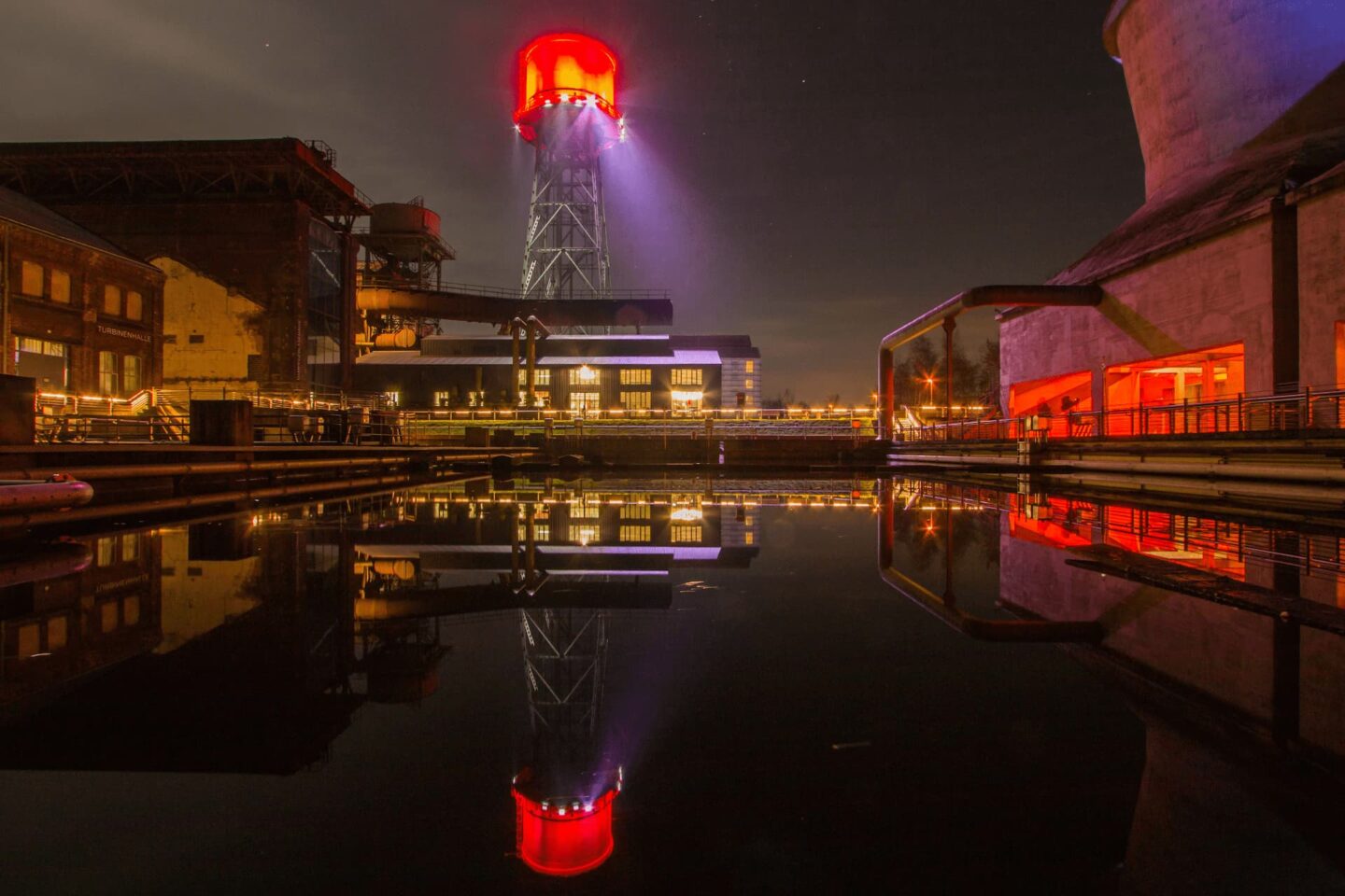 Auf einem Industriegelände in Bochum ist nachts eine hohe, rot beleuchtete Struktur zu sehen, deren Spitze Spiegelbilder auf die ruhige Wasseroberfläche darunter wirft. Gebäude und andere Industrieanlagen umgeben das Gelände, alle mit warmem Licht beleuchtet, das einen Kontrast zum dunklen Himmel bildet.