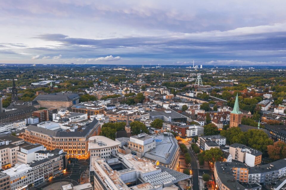 Luftaufnahme der Stadtlandschaft von Bochum in der Abenddämmerung, mit einer Mischung aus moderner und historischer Architektur, Grünflächen mit Bäumen und Straßen mit wenig Verkehr. Der Himmel ist mit Streifen blauer und grauer Wolken bedeckt.